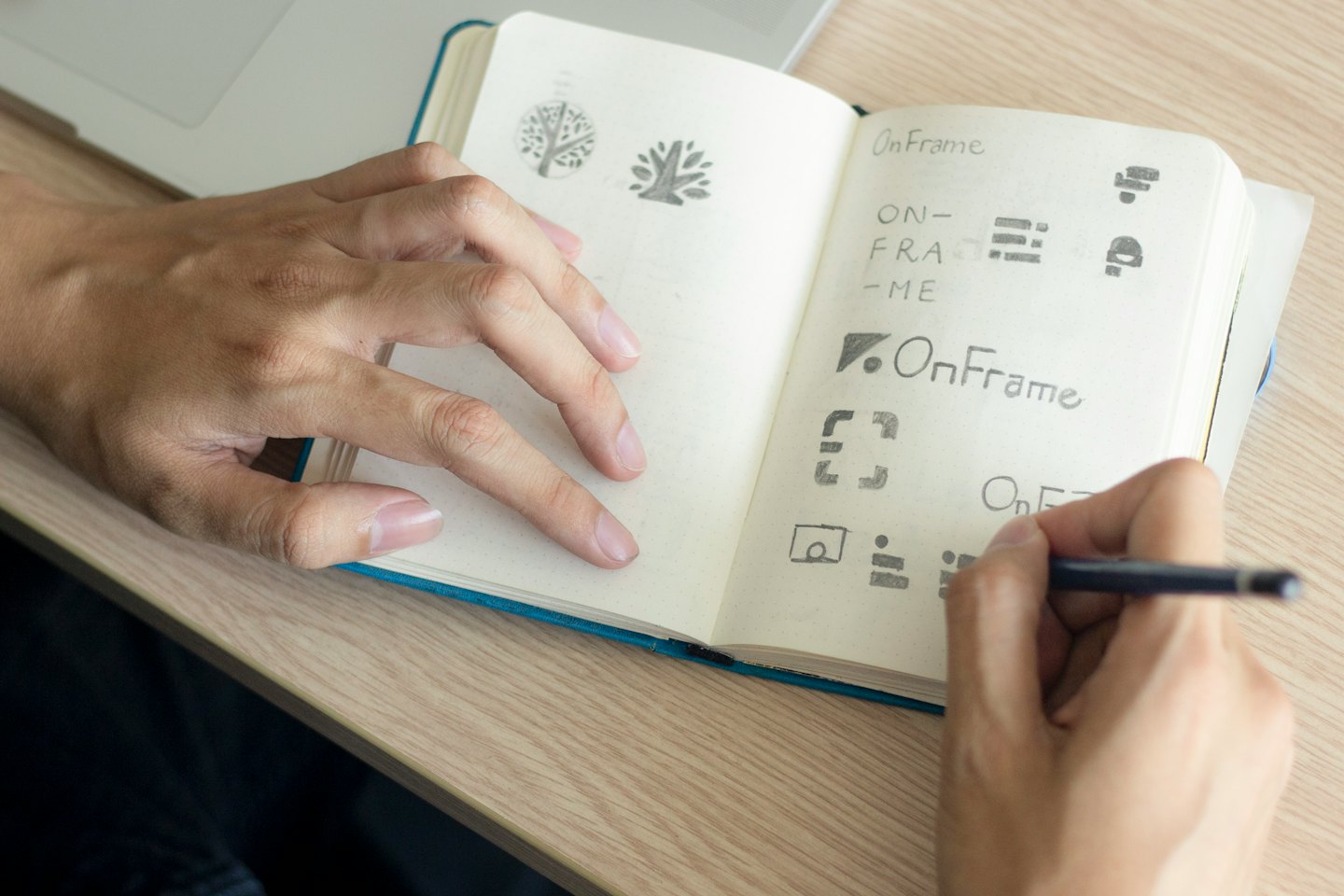 Hands sketching logo designs in a notebook on a desk.